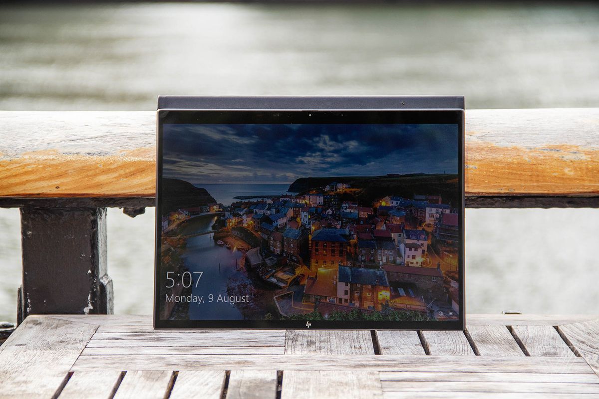 HP Elite Folio in tablet mode leaning against a railing and a river in the background on a wooden table.  The screen displays a Lock screen with a twilight city along the river.