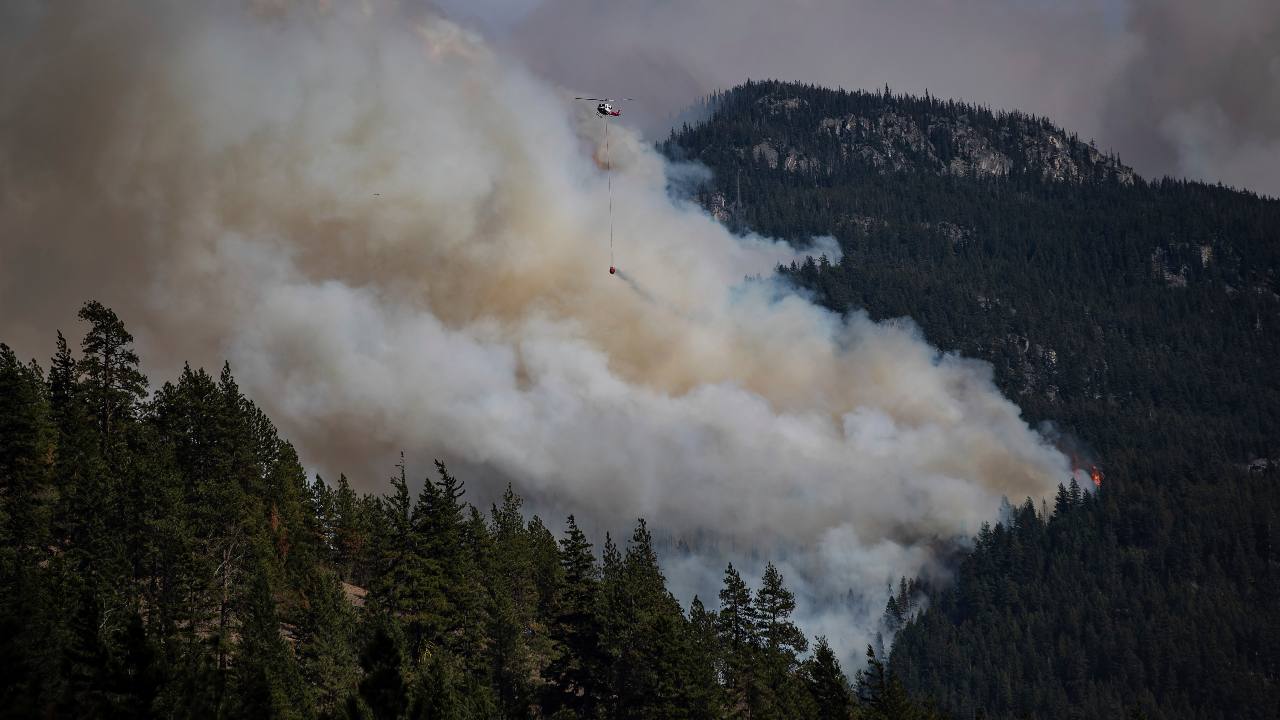 A helicopter with a water tank flies past a forest fire at Lytton Creek, which returns in the mountains near Lytton, British Columbia, on Sunday, August 15, 2021. (via Darryl Dyck / The Canadian Press AP)