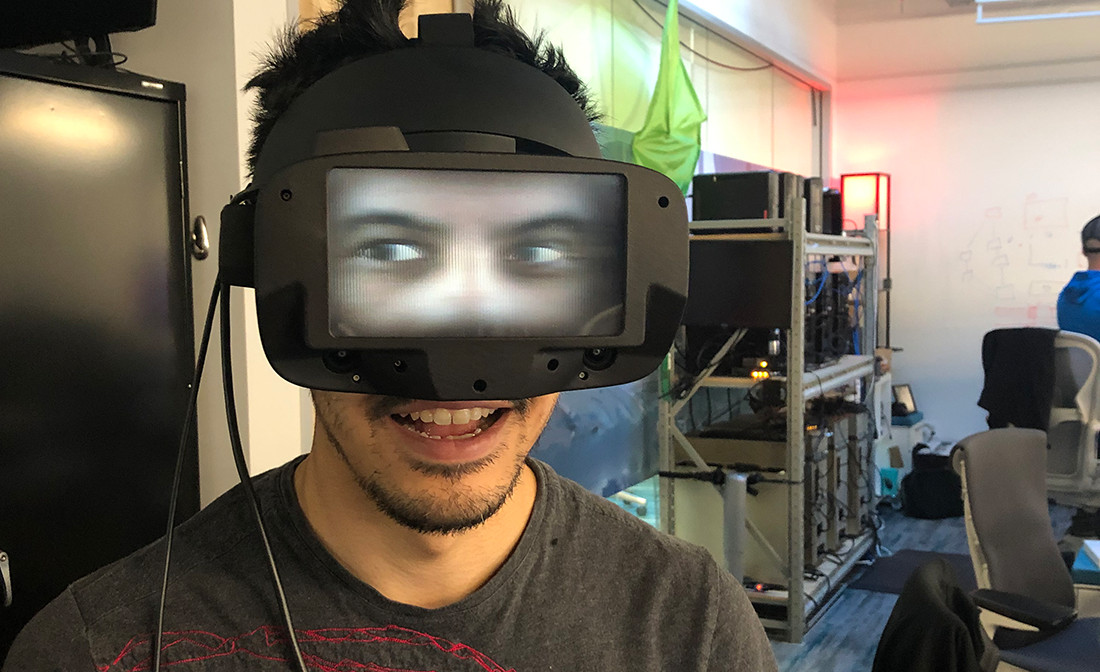 FRL researcher Nathan Matsuda wears custom Oculus Rift S headphones with a flat 3D screen on the front and looks above him.